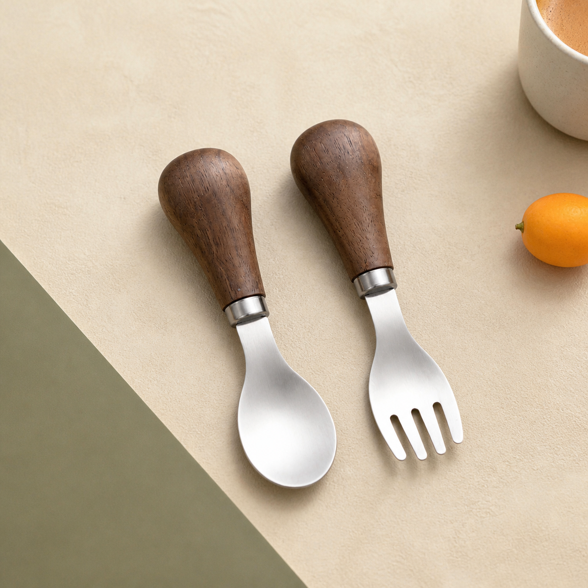 Petite spoon and four-tine fork with rounded walnut handles on a warm neutral and olive background.