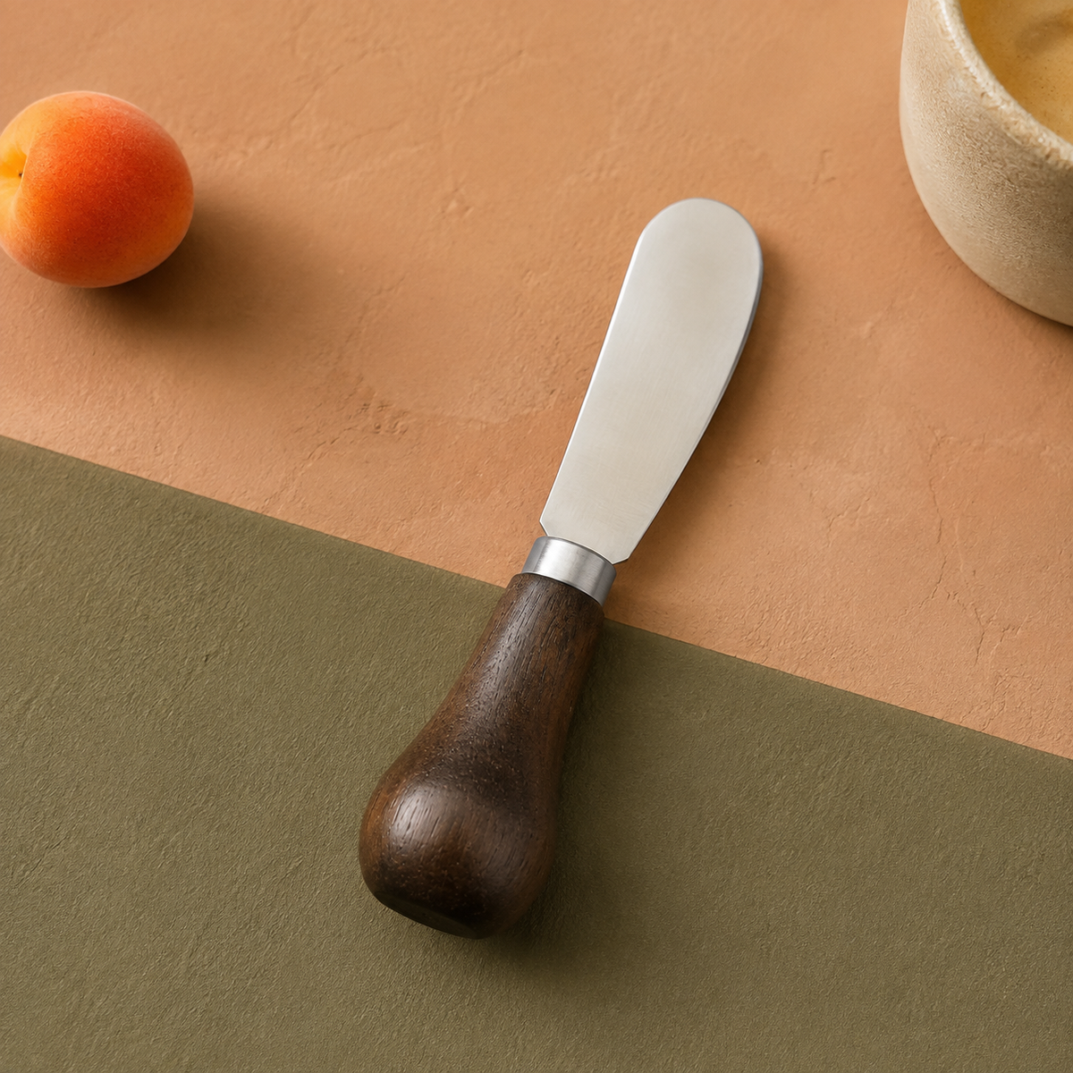 Compact walnut-handled butter spreader on a terracotta and muted olive surface.
