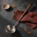Long walnut-handled stainless steel soup ladle arranged on a dark blue-grey surface with rust-coloured linen.