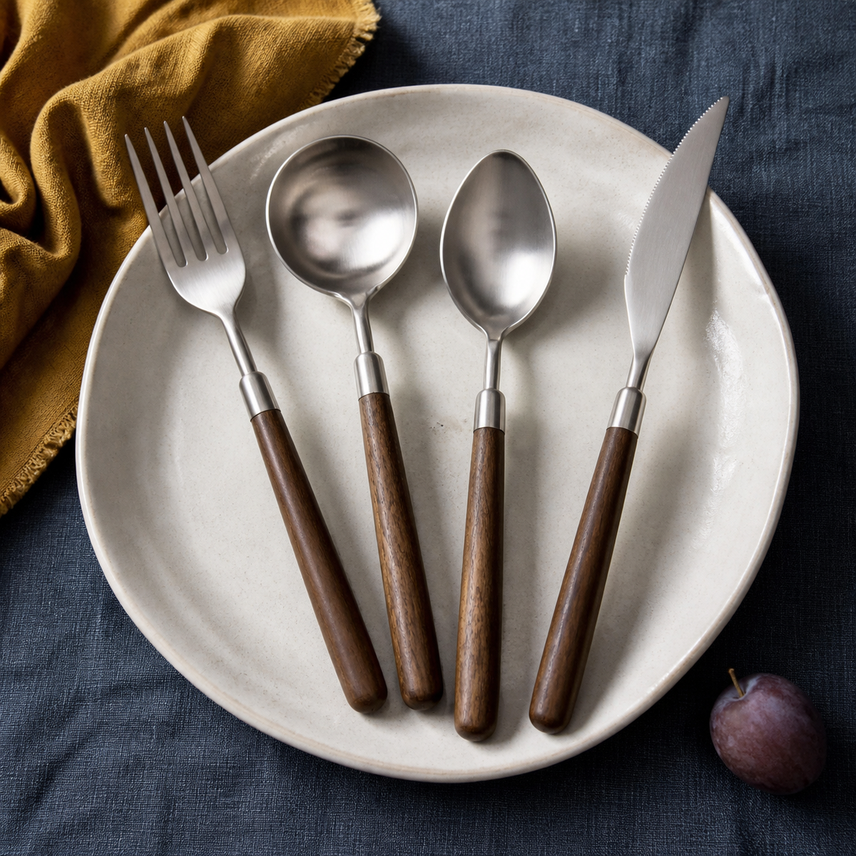 Four-piece walnut-handled stainless steel flatware set arranged on an ivory plate over blue-grey linen.