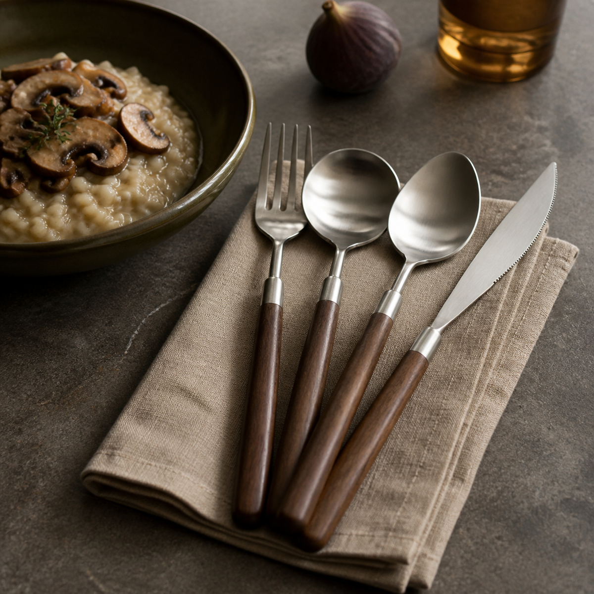Four-piece walnut-handled flatware set resting on an oat linen napkin beside a mushroom risotto bowl.